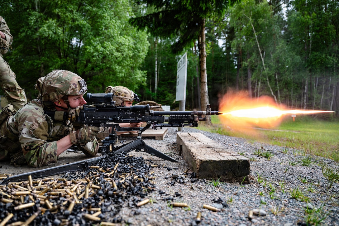 Ammo sits on the ground next to soldiers as they fire weapons in a wooded area, creating an orange fireball.