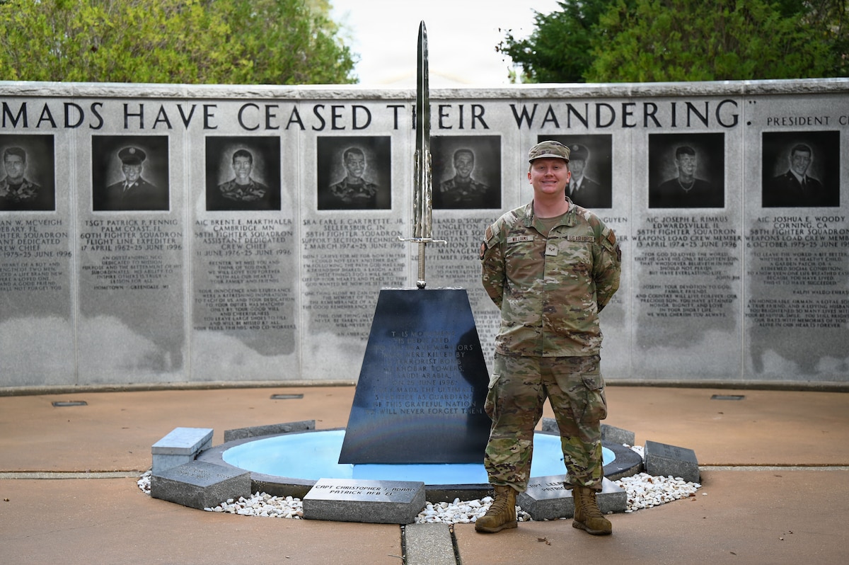 A man wearing a camouflage military uniform stands in front of a sword wedged into a stone block. Behind him is a wall memorializing the fallen service members of a tower bombing.