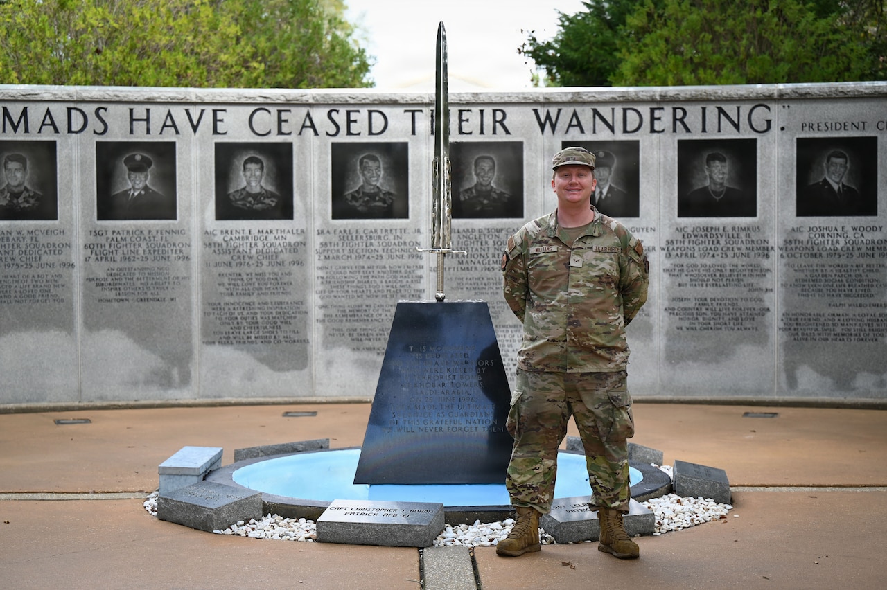 A man wearing a camouflage military uniform stands in front of a sword wedged into a stone block. Behind him is a wall memorializing the fallen service members of a tower bombing.