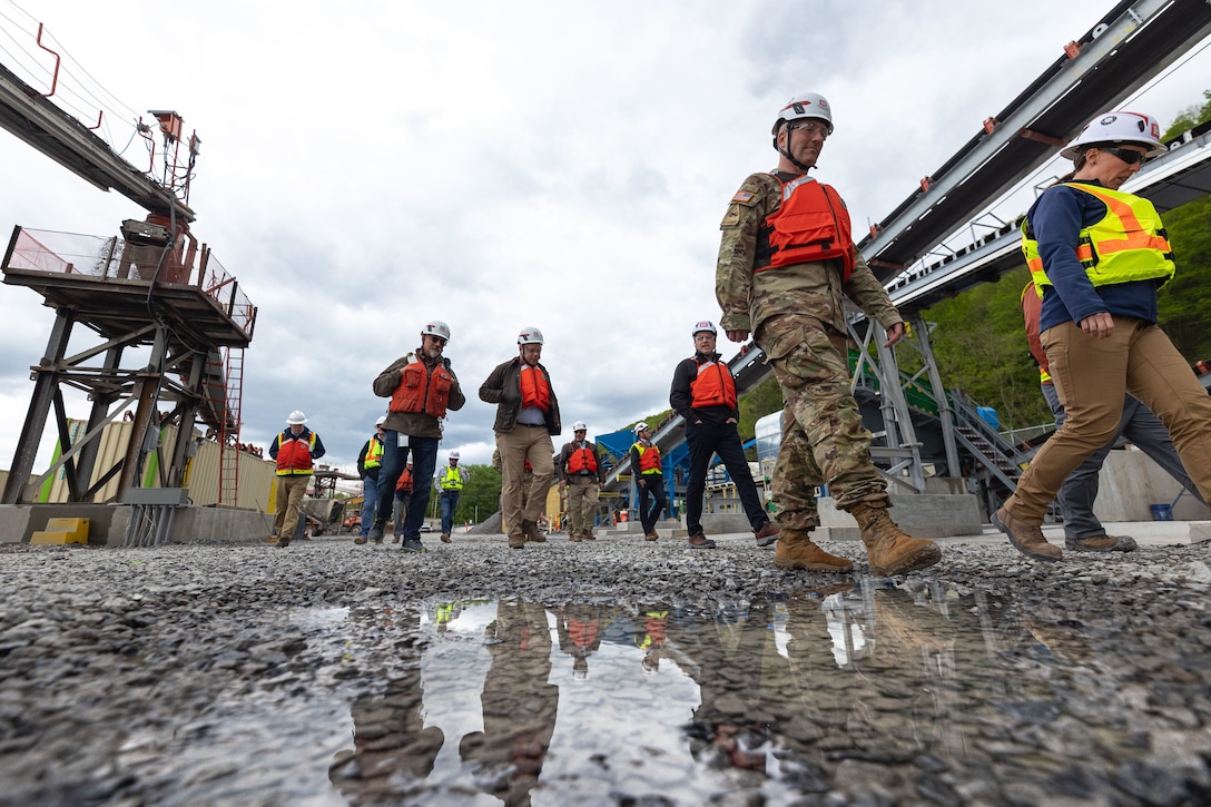 A group of people in hard hats and life vests walk along a gravel road at a construction site. A puddle in the foreground reflects a soldier in uniform.