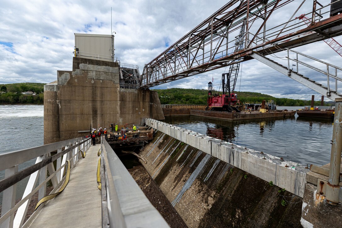 A wide view of a dam, with several workers in hard hats on a walkway overlooking the water where a barge with a large crane is docked.
