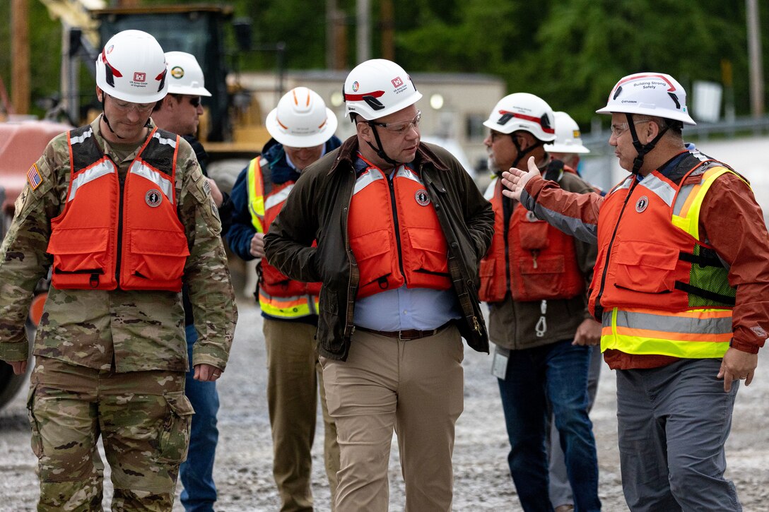 A group of men in hard hats and life vests converse on a gravel lot at a construction site.