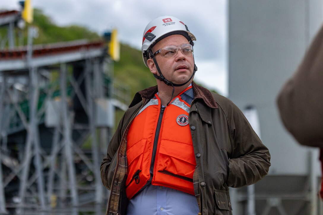 A man wearing a hard hat, glasses, and an orange life vest over a brown jacket looks to the side at a construction site.