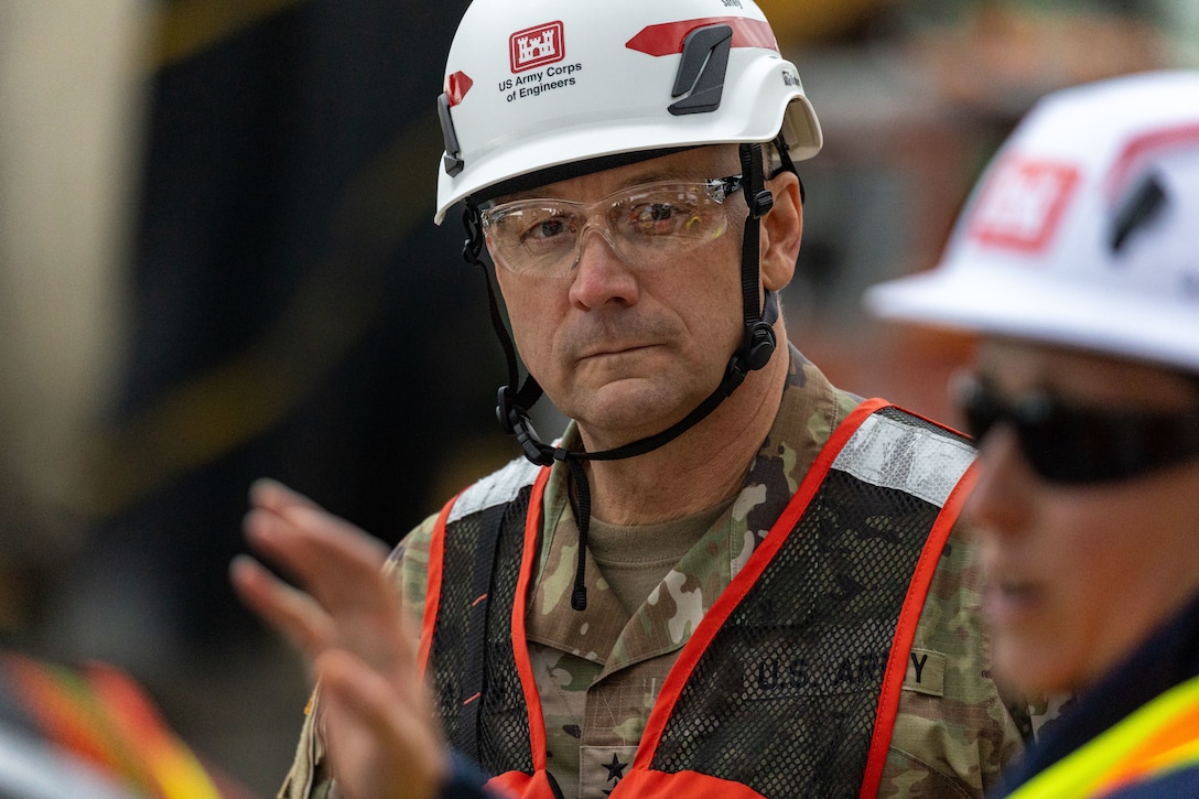 A close-up of a man in a military uniform and a "US Army Corps of Engineers" hard hat, looking seriously at someone off-camera.