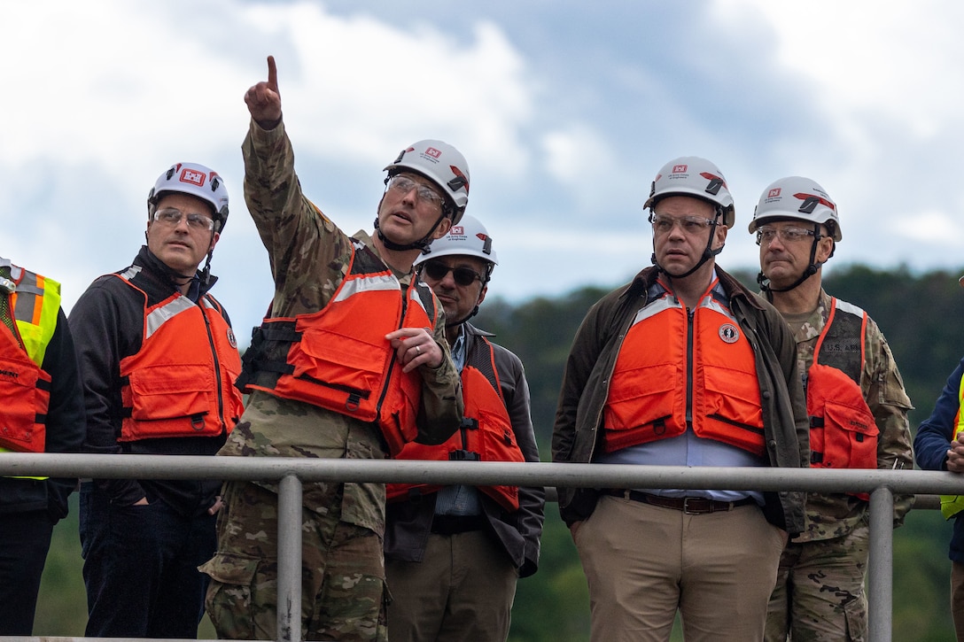 A group of men in hard hats and orange life vests look up as one man in a military uniform points toward the sky.