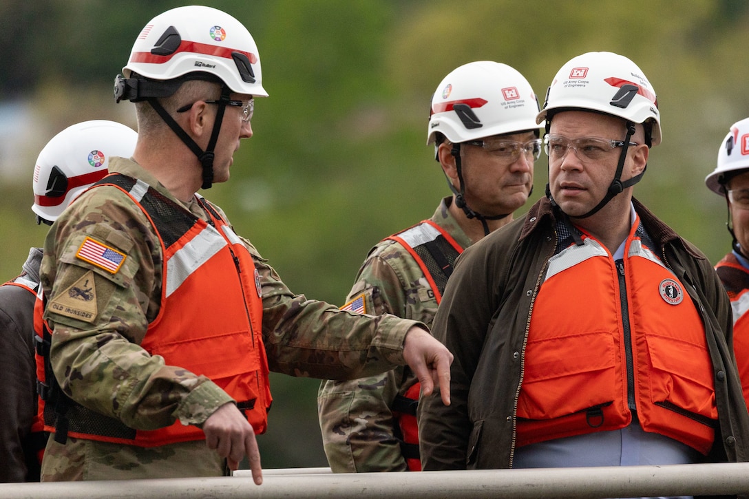 A man in a military uniform with an "Old Ironsides" patch gestures while speaking to another man in a hard hat and life vest.
