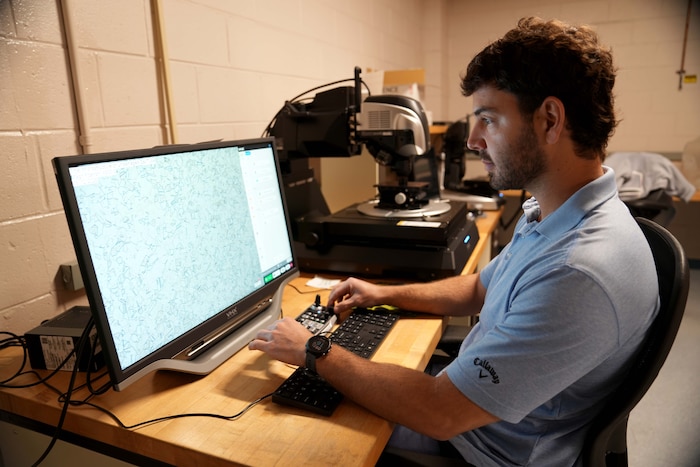 Photo shows man using devices on a small part and looking at a large digital screen
