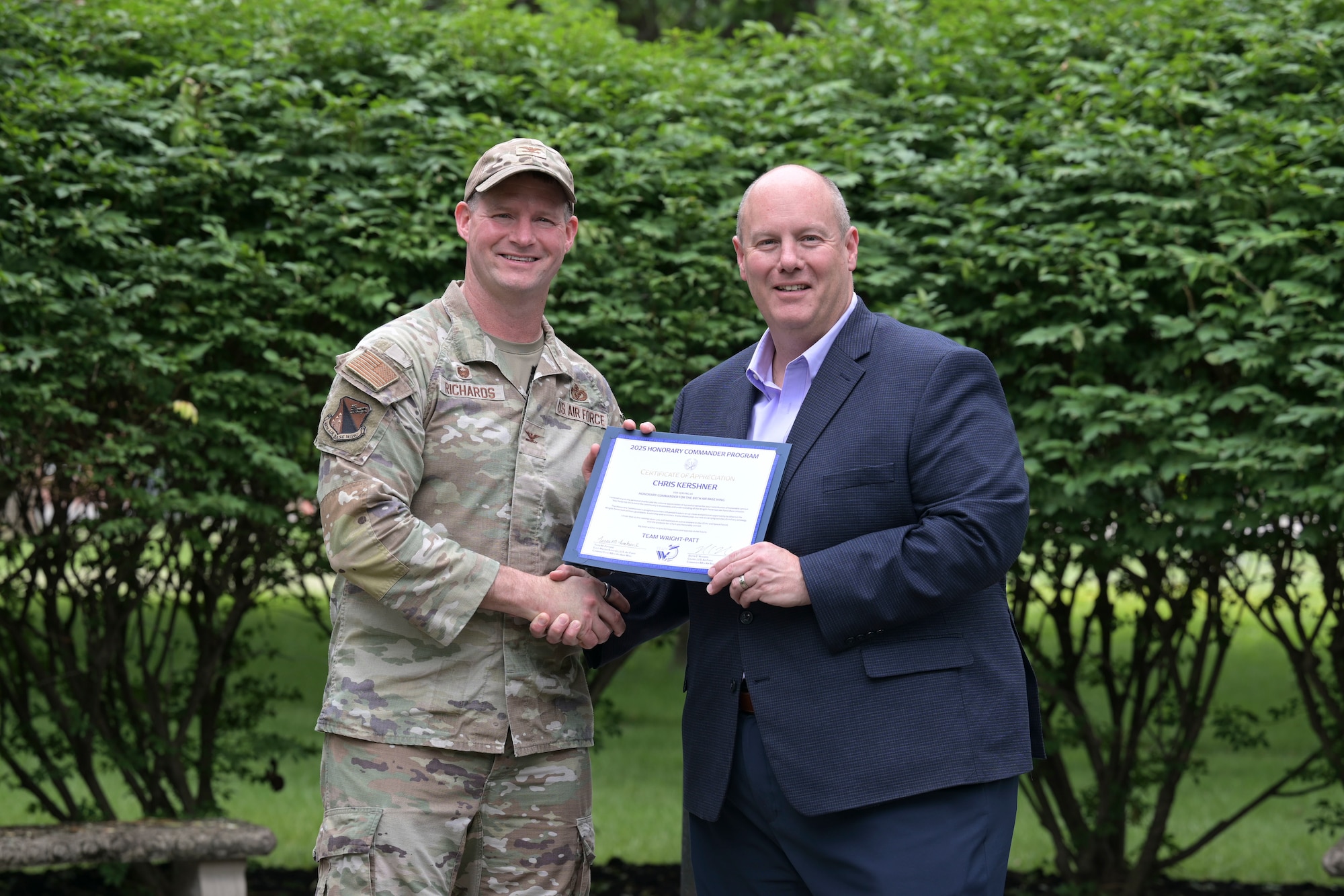 A man in a camouflage uniform and ball cap presents a certificate to a man in a navy blue suit