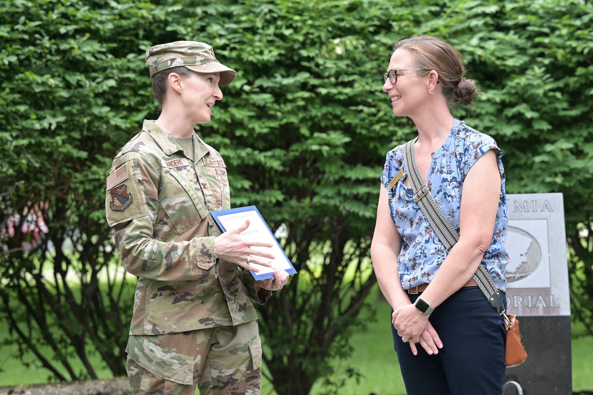 A woman in a camouflage uniform smiles as she presents a certificate to another woman wearing civilian attire
