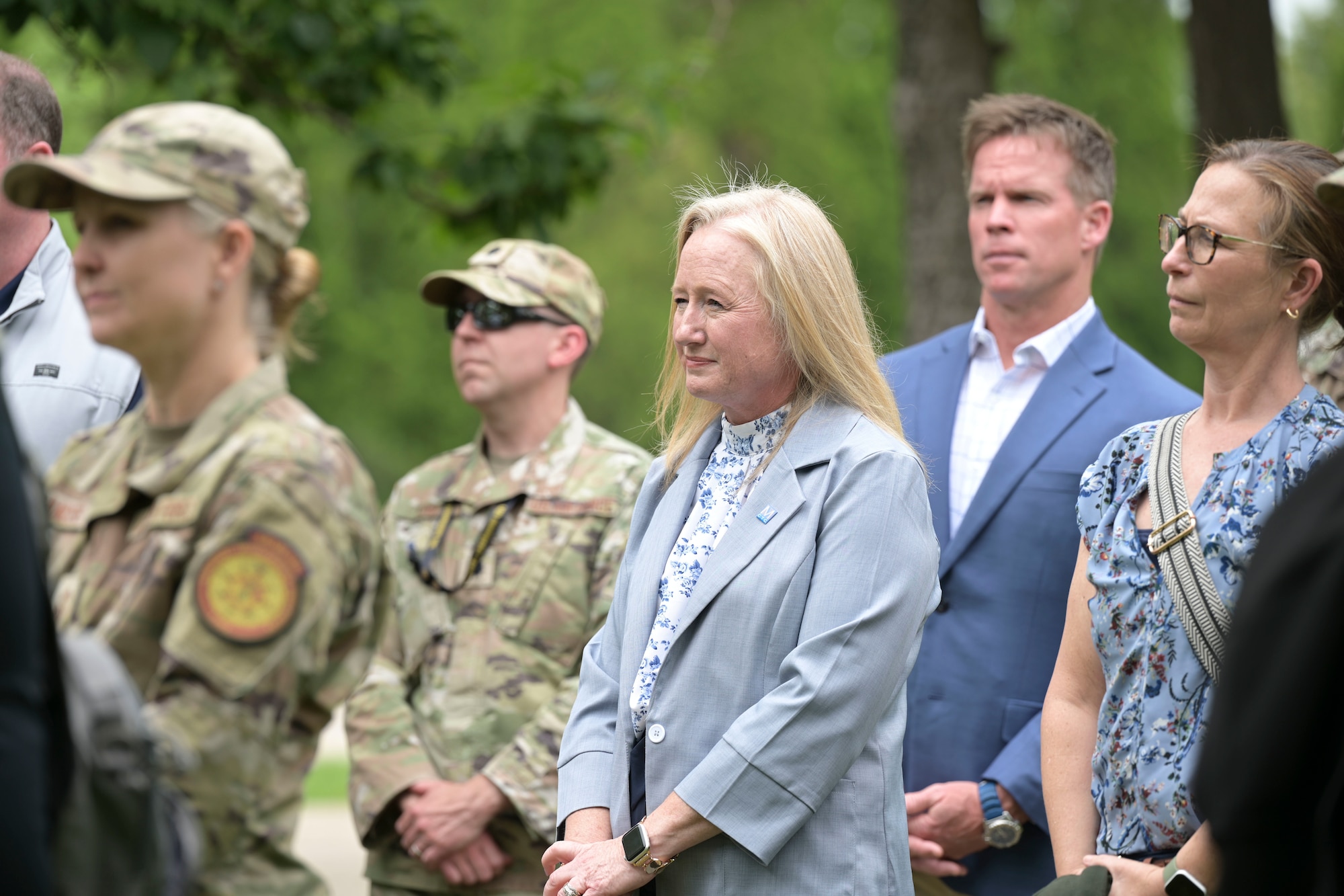 A group of men and women in civilian attire and military uniforms stand while listening to opening remarks outside