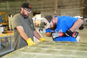 Photo shows two men using a handheld cutter on a large piece of metal