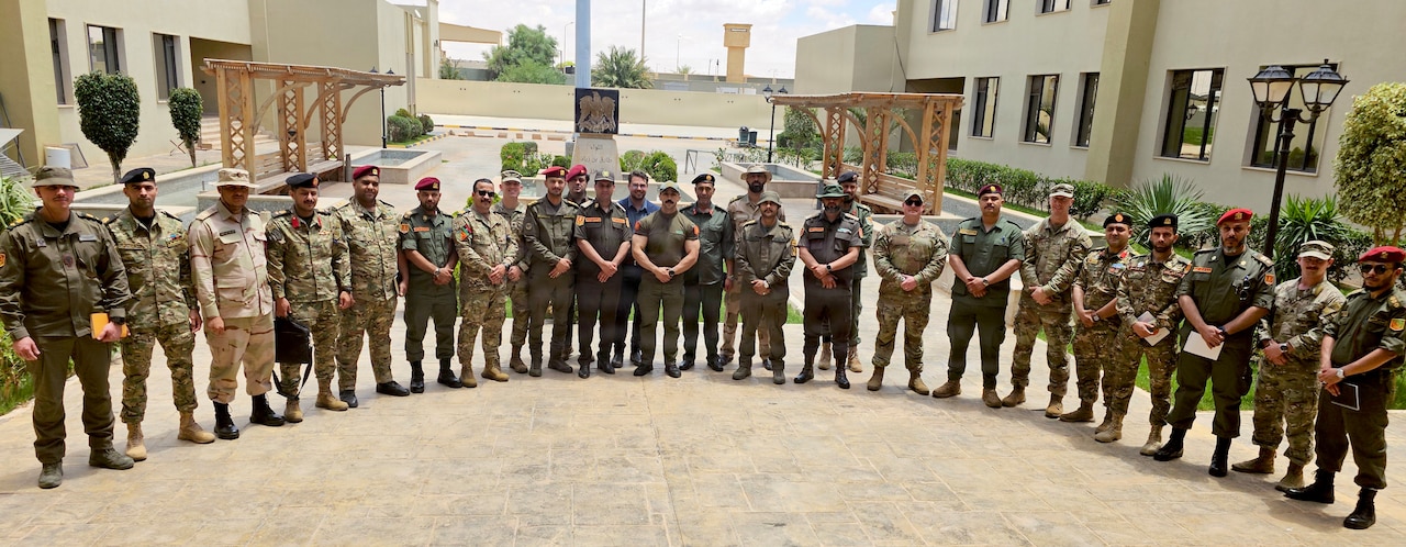 Two dozen men in military uniforms pose for a group photo outside in a courtyard.