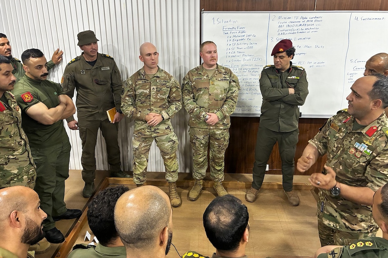 A dozen men in military uniforms stand in a circle watching another man in similar attire speak inside a classroom.