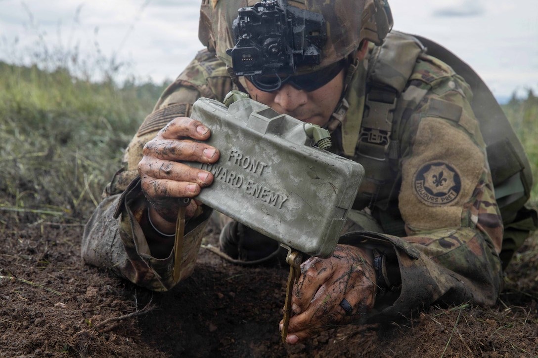 A soldier in tactical gear plants a mine in the dirt against a gray sky.
