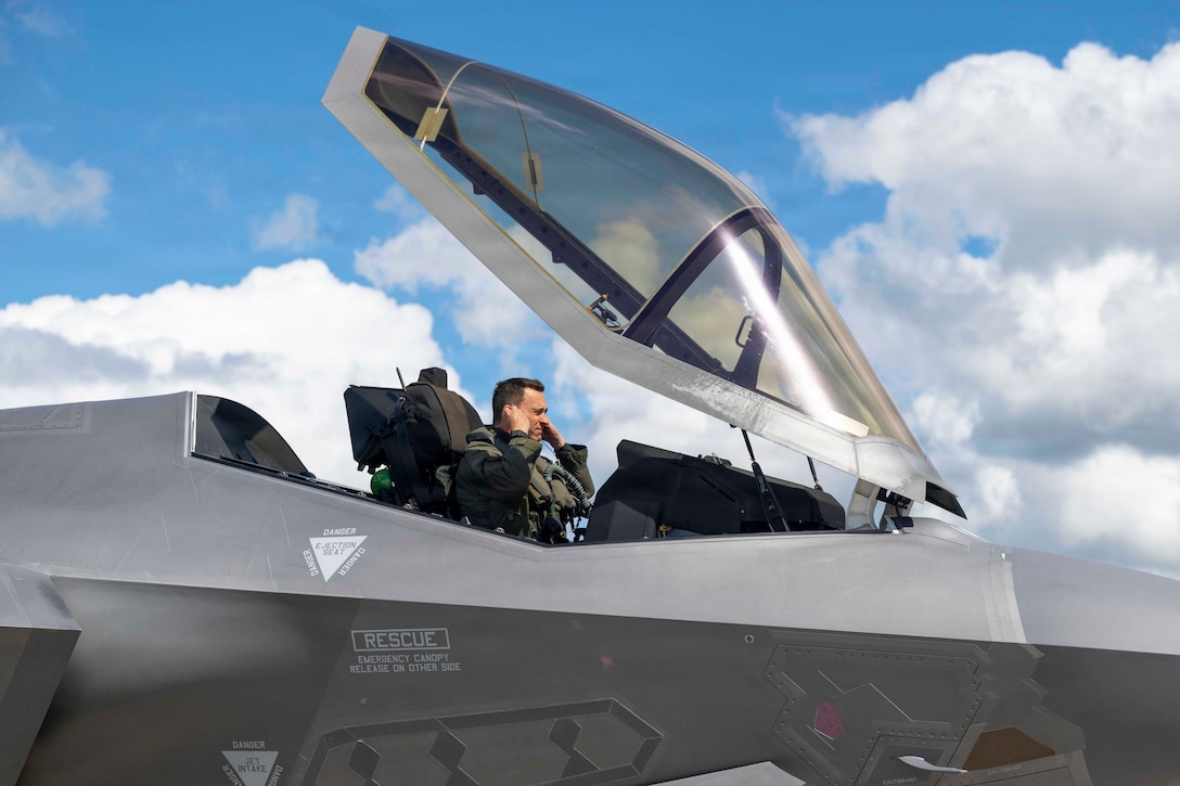 A close-up of an airman putting in earpieces while sitting in an open cockpit against a blue sky.