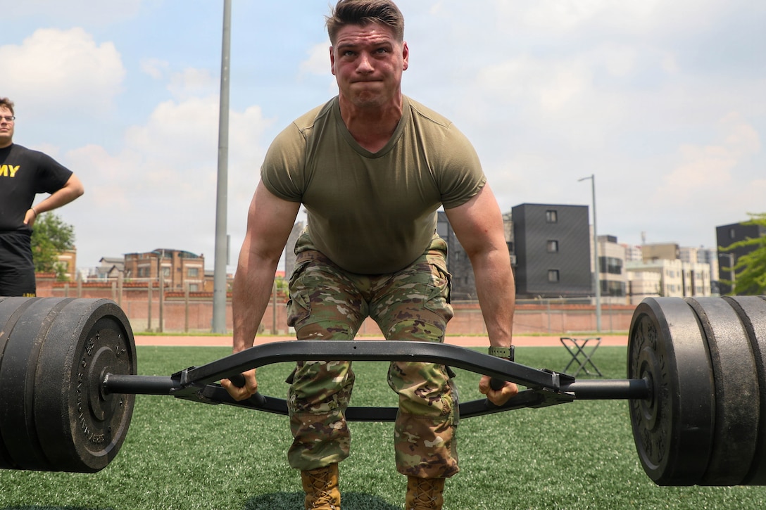 A soldier strains while deadlifting hundreds of pounds on a turf during the day.