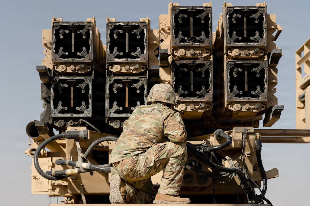 A soldier kneels in front of a military vehicle against a grayish blue sky.