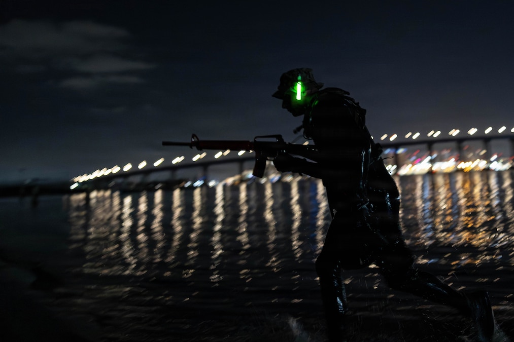 A Marine holds a weapon while running near a body of water in the dark illuminated by a green glowstick and bridge lights in the background.