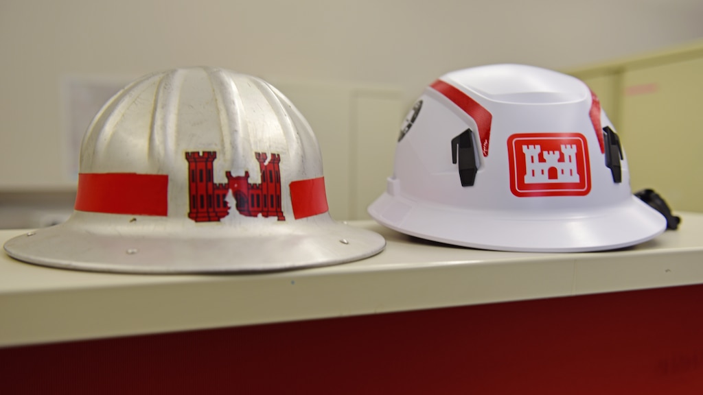 The white U.S. Army Corps of Engineers hard hat on the right with red stripes and castle logo is very distinctive and recognizable on any construction or dam project site. This modern hard hat comes with a chin strap and has an internal suspension that stretches and deforms, absorbs kinetic energy and distributes the force evenly. The rigid shell prevents sharp, heavy, or high-velocity objects such as falling bolts or tools from piercing the helmet. Between 1930 and 1950 the metal hard hat on the left provided USACE employees protection but its conductivity made it dangerous near electrical hazards. Hard Hat Awareness Week is June 8-14, 2026. (USACE Photo by Lee Roberts)