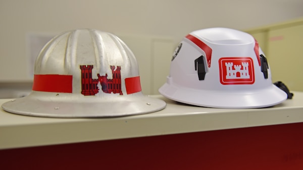 The white U.S. Army Corps of Engineers hard hat on the right with red stripes and castle logo is very distinctive and recognizable on any construction or dam project site. This modern hard hat comes with a chin strap and has an internal suspension that stretches and deforms, absorbs kinetic energy and distributes the force evenly. The rigid shell prevents sharp, heavy, or high-velocity objects such as falling bolts or tools from piercing the helmet. Between 1930 and 1950 the metal hard hat on the left provided USACE employees protection but its conductivity made it dangerous near electrical hazards. Hard Hat Awareness Week is June 8-14, 2026. (USACE Photo by Lee Roberts)