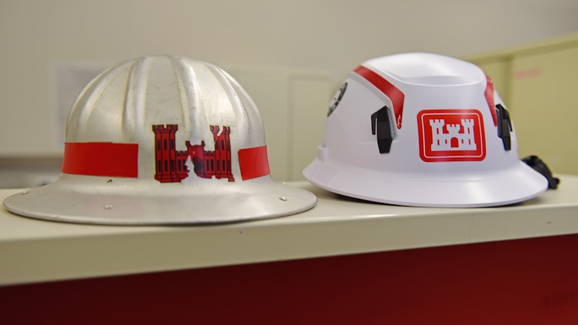 The white U.S. Army Corps of Engineers hard hat on the right with red stripes and castle logo is very distinctive and recognizable on any construction or dam project site. This modern hard hat comes with a chin strap and has an internal suspension that stretches and deforms, absorbs kinetic energy and distributes the force evenly. The rigid shell prevents sharp, heavy, or high-velocity objects such as falling bolts or tools from piercing the helmet. Between 1930 and 1950 the metal hard hat on the left provided USACE employees protection but its conductivity made it dangerous near electrical hazards. Hard Hat Awareness Week is June 8-14, 2026. (USACE Photo by Lee Roberts)