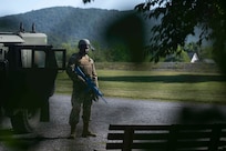 An Airman stands in tactical gear holding a training weapon in front of large hills with the silhouette of leaves between the viewer and the Airman.