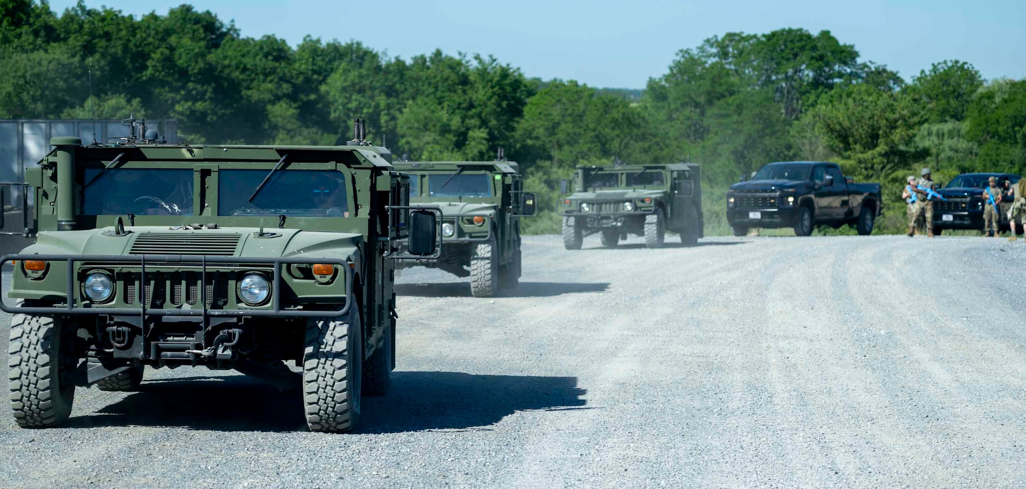 A line of Humvees and trucks approaches down a gravel road.