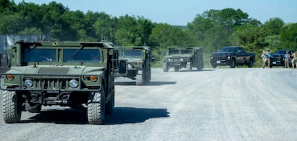A line of Humvees and trucks approaches down a gravel road.