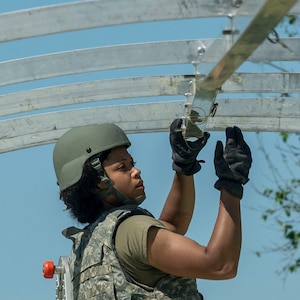 An Airman wearing tactical gear reaches up to assemble metal tent pieces.