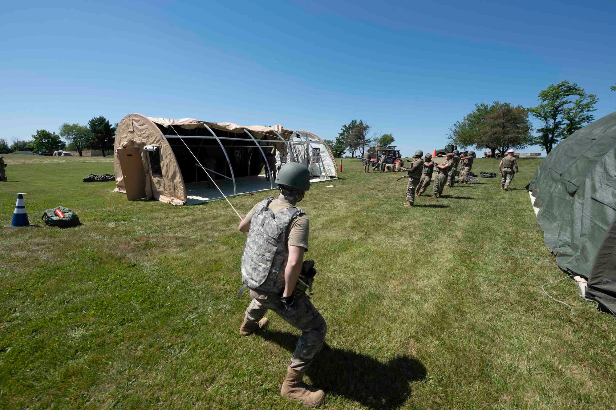 Airmen wearing helmets and vests pull covers over tent frames with ropes.