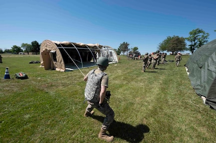 Airmen wearing helmets and vests pull covers over tent frames with ropes.