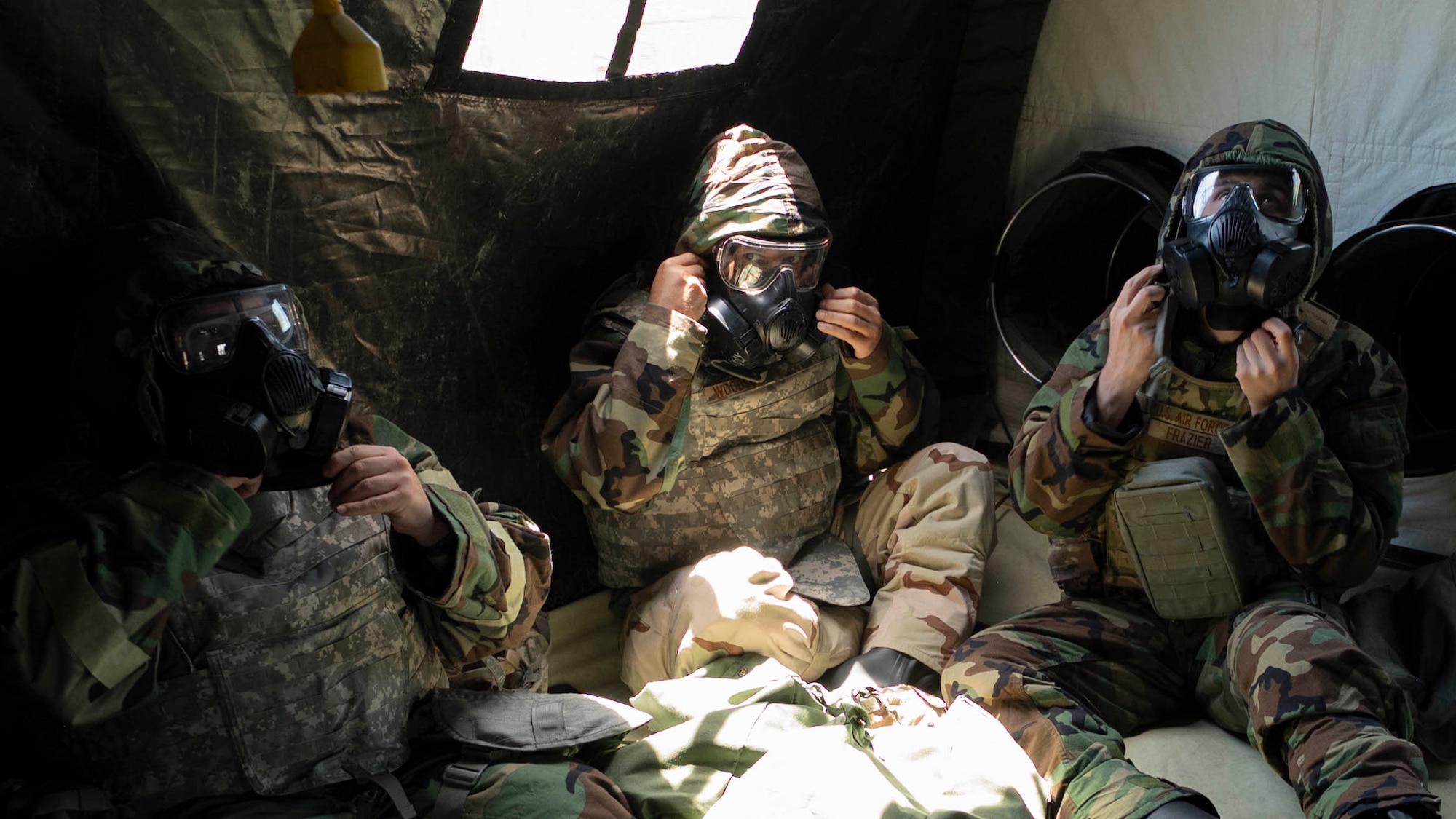 Three Airmen sit in a tent tightening gas masks.