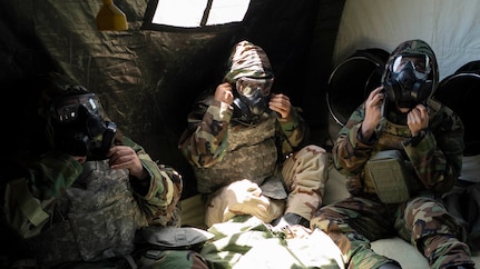 Three Airmen sit in a tent tightening gas masks.