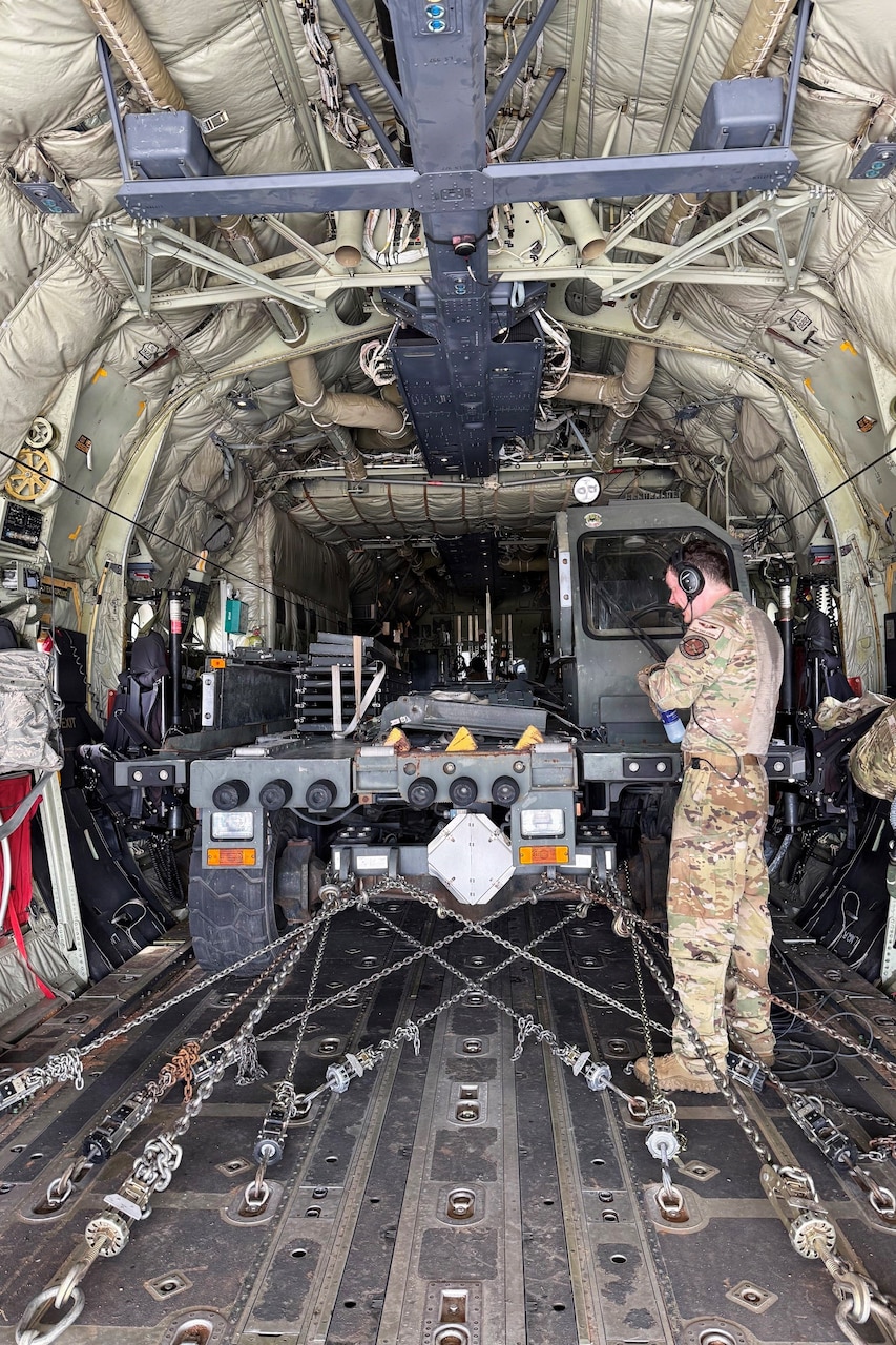 A man wearing a camouflage military uniform stands in the back of a military aircraft while looking at a military loading vehicle.