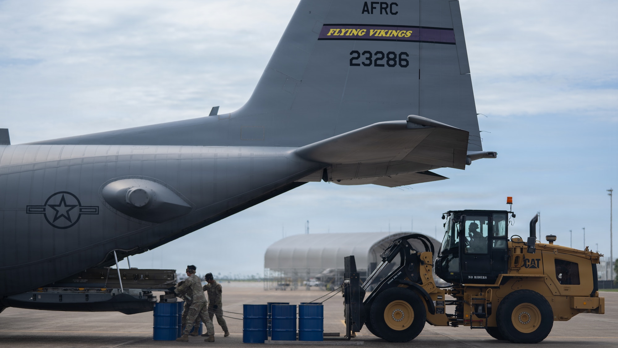 2 people stand behind a military aircraft unloading cargo, while a third person is behind them in a loading vehicle