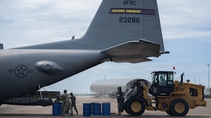 2 people stand behind a military aircraft unloading cargo, while a third person is behind them in a loading vehicle