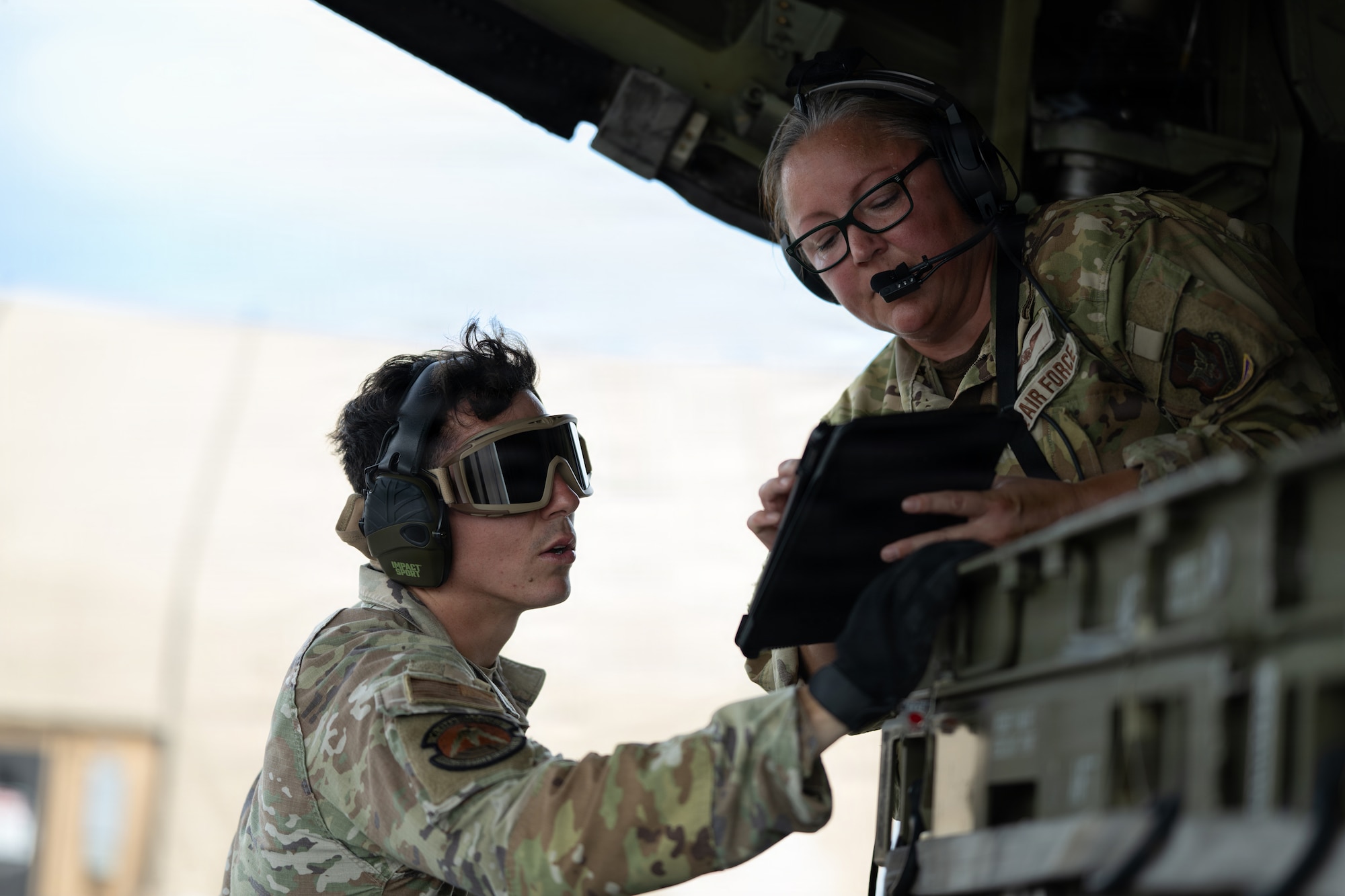 Two people wearing military uniforms talk and point at a checklist while standing in the back of a military aircraft