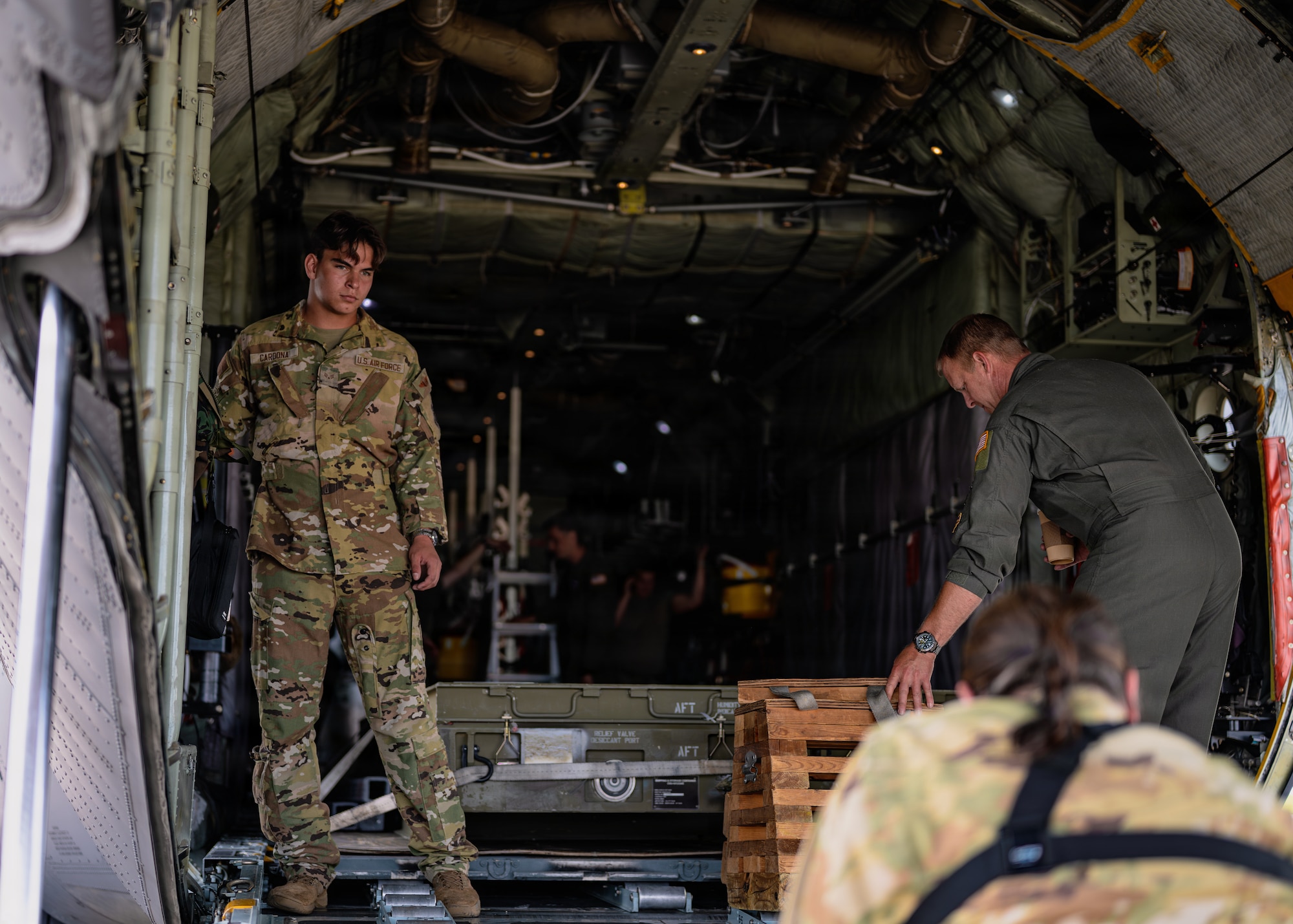 Three military members stand in the back of a C-130 aircraft. One of them appears to be reaching for a piece of cargo, while the other two are looking at something out of frame