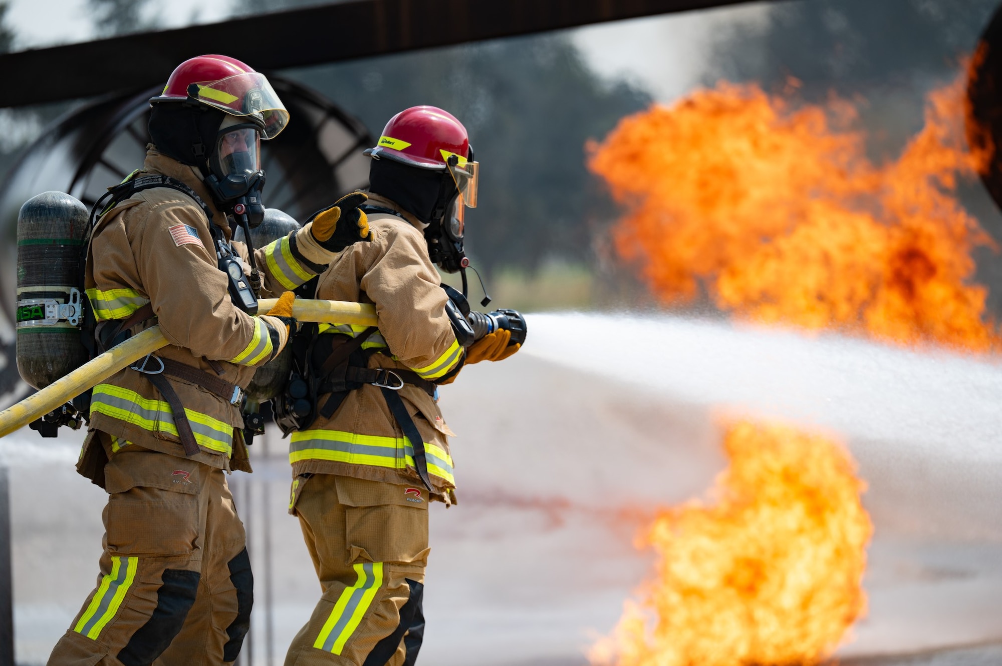 Two men in firefighter gear put out a fire with a hose spraying water.