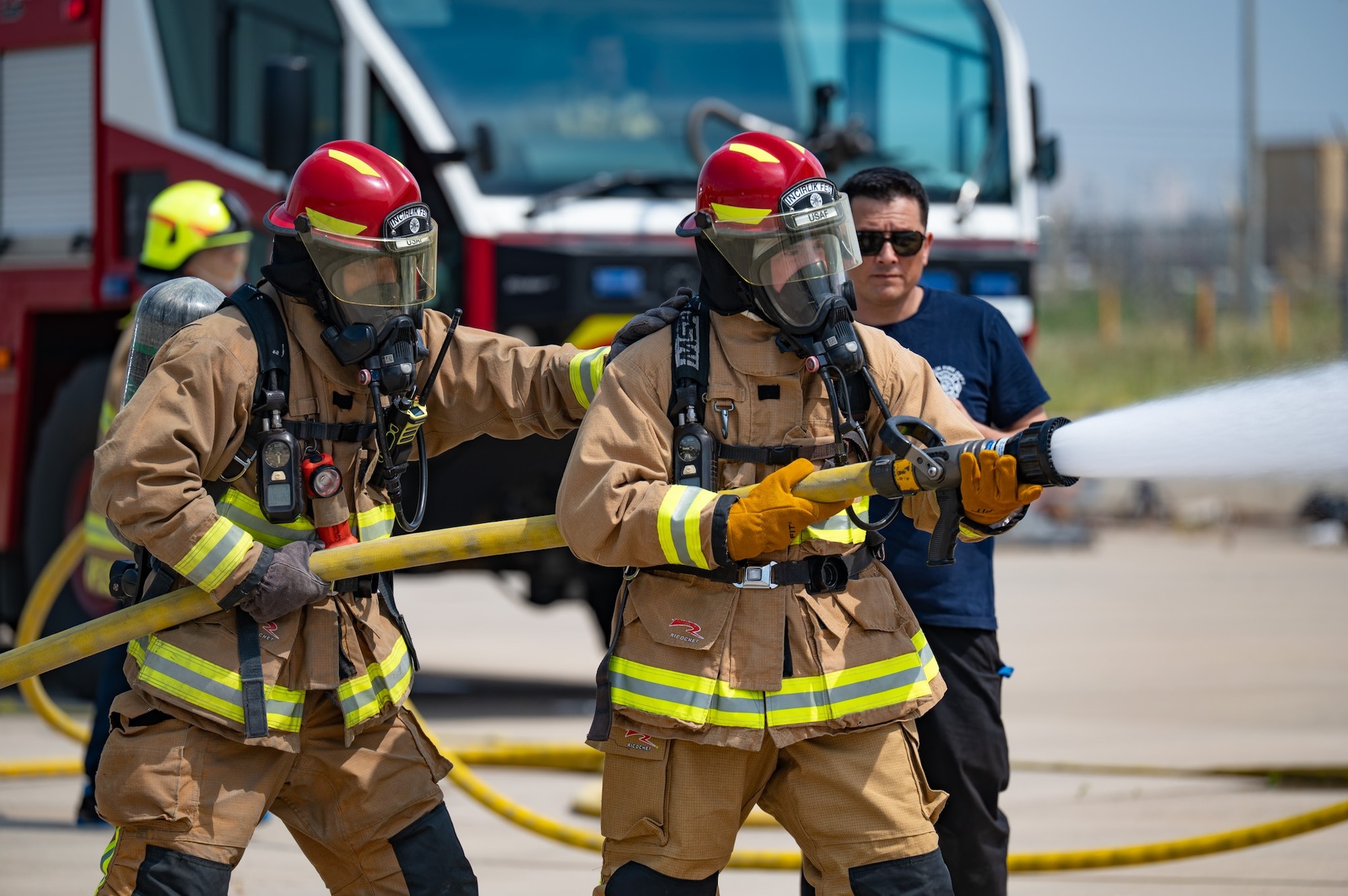 Two men in firefighter gear hold a firefighter hose that as it sprays water.