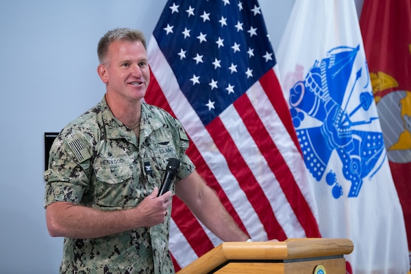 A man wearing a camouflage military uniform stands behind a podium speaking into a microphone.