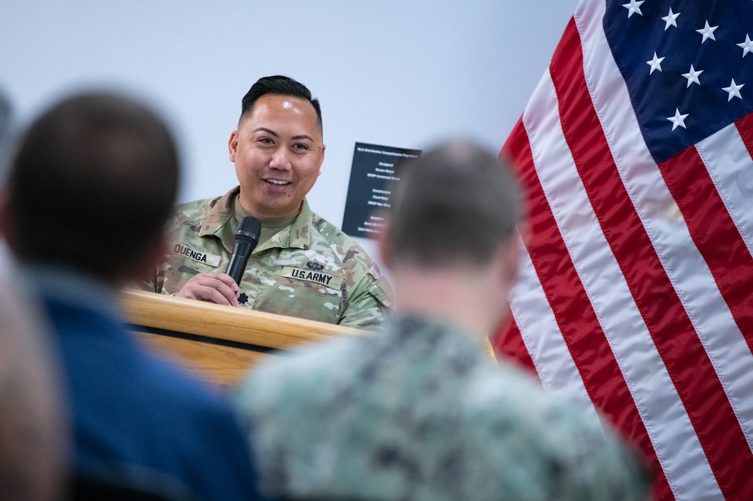 A man wearing a camouflage military uniform stands behind a podium speaking into a microphone.