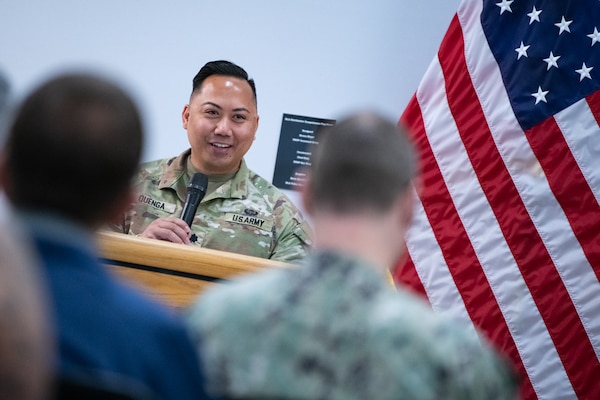 A man wearing a camouflage military uniform stands behind a podium speaking into a microphone.