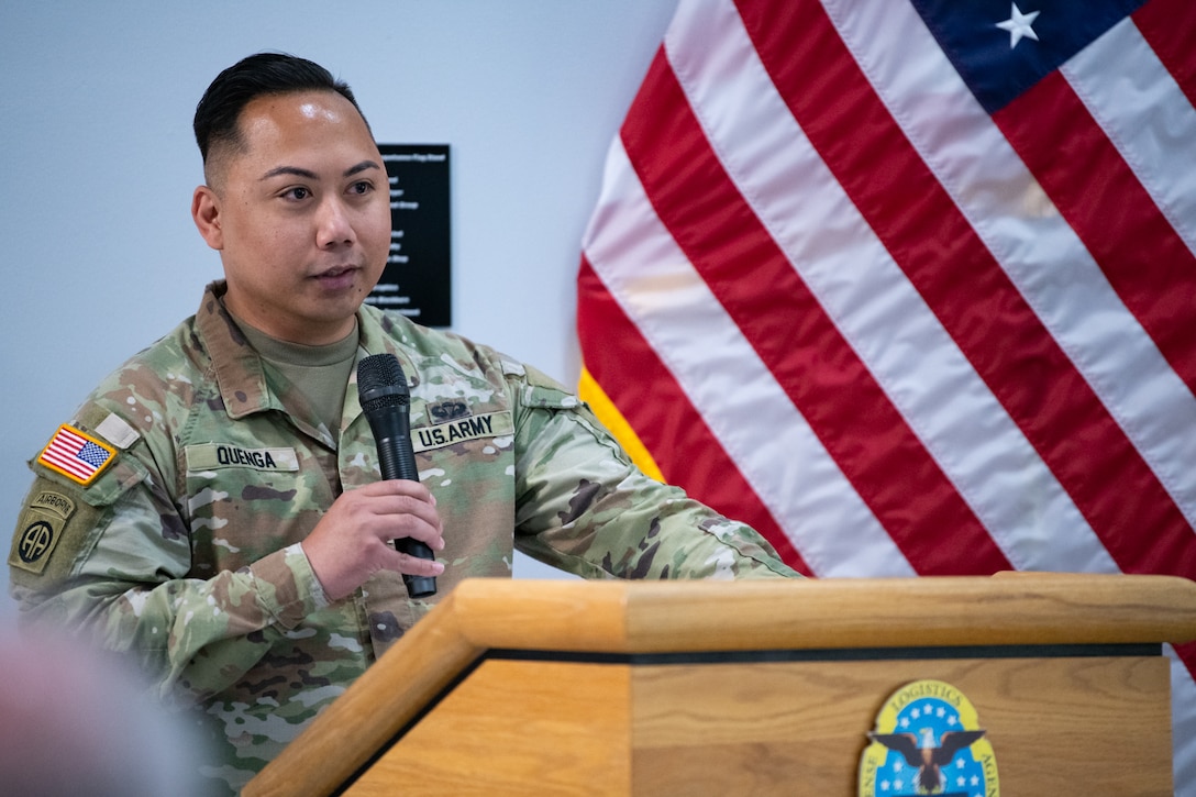 A man wearing a camouflage military uniform stands behind a podium speaking into a microphone.