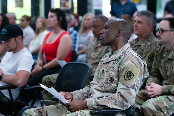 A group of people, several wearing camouflage military uniforms, sit in an audience in a building lobby.