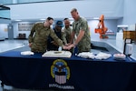 Four men, three of them wearing camouflage military uniforms, stand behind a table with a cake on it.