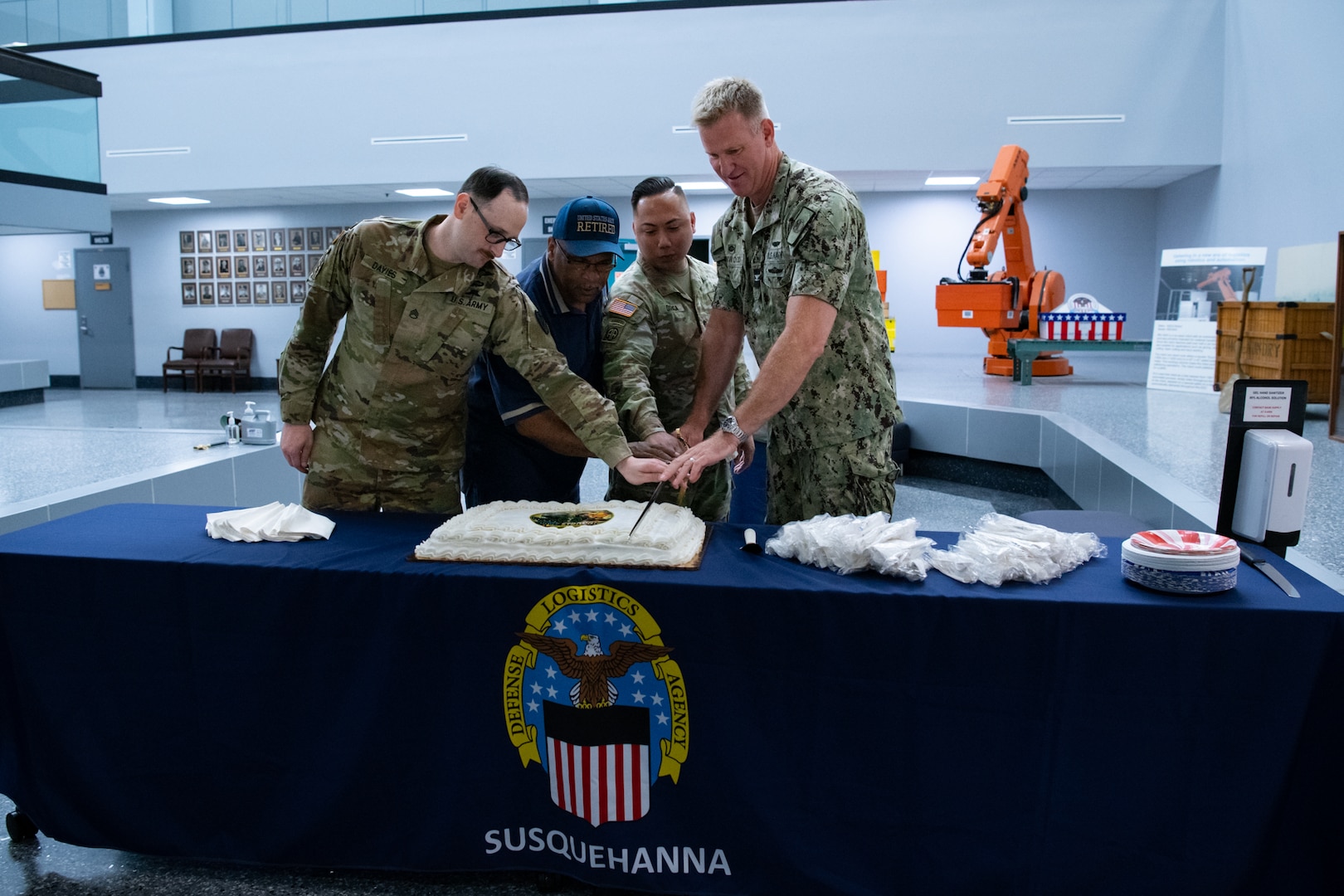 Four men, three of them wearing camouflage military uniforms, stand behind a table with a cake on it.
