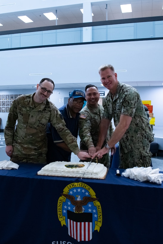 Four men, three of them wearing camouflage military uniforms, stand behind a table with a cake on it.