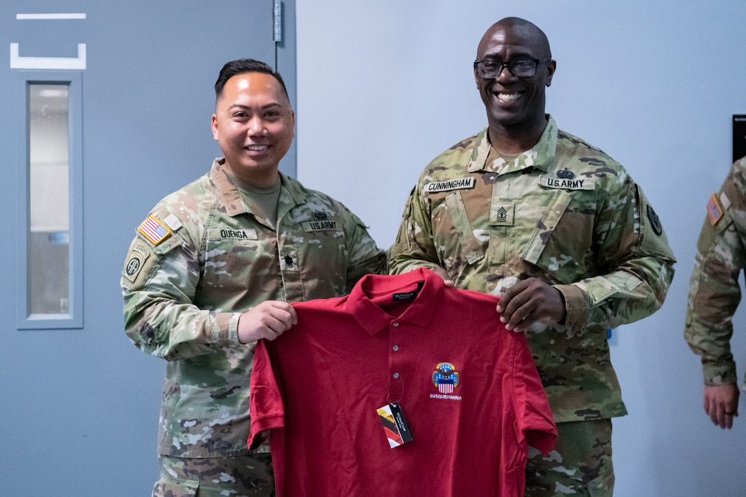 Two men wearing camouflage military uniforms stand together holding a red shirt.