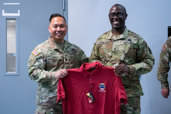 Two men wearing camouflage military uniforms stand together holding a red shirt.